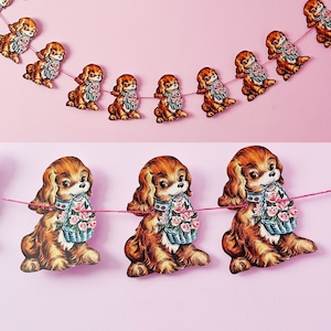 May include: A garland of brown and white paper cutouts of dogs holding baskets of pink flowers. The garland is strung with pink string and is hanging against a pink background.