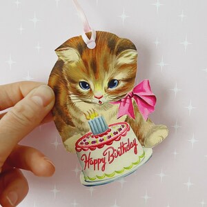 May include: A vintage birthday card featuring a cute cartoon cat holding a pink and white birthday cake with the words "Happy Birthday" on it.