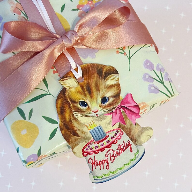 May include: A gift tag featuring a cartoon cat with a pink bow holding a birthday cake with the text "Happy Birthday". The tag is tied with a pink ribbon to a white and green floral gift wrap.