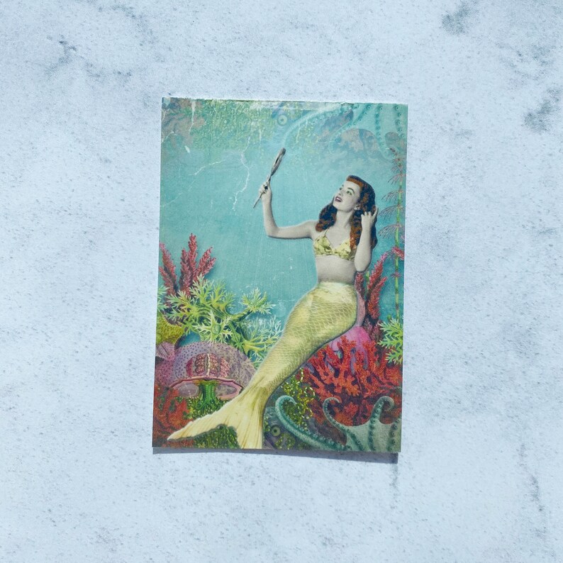 May include: A vintage postcard featuring a mermaid with red hair sitting on a large seashell with an octopus and coral surrounding her. The mermaid is holding a comb in her right hand and looking to the left.