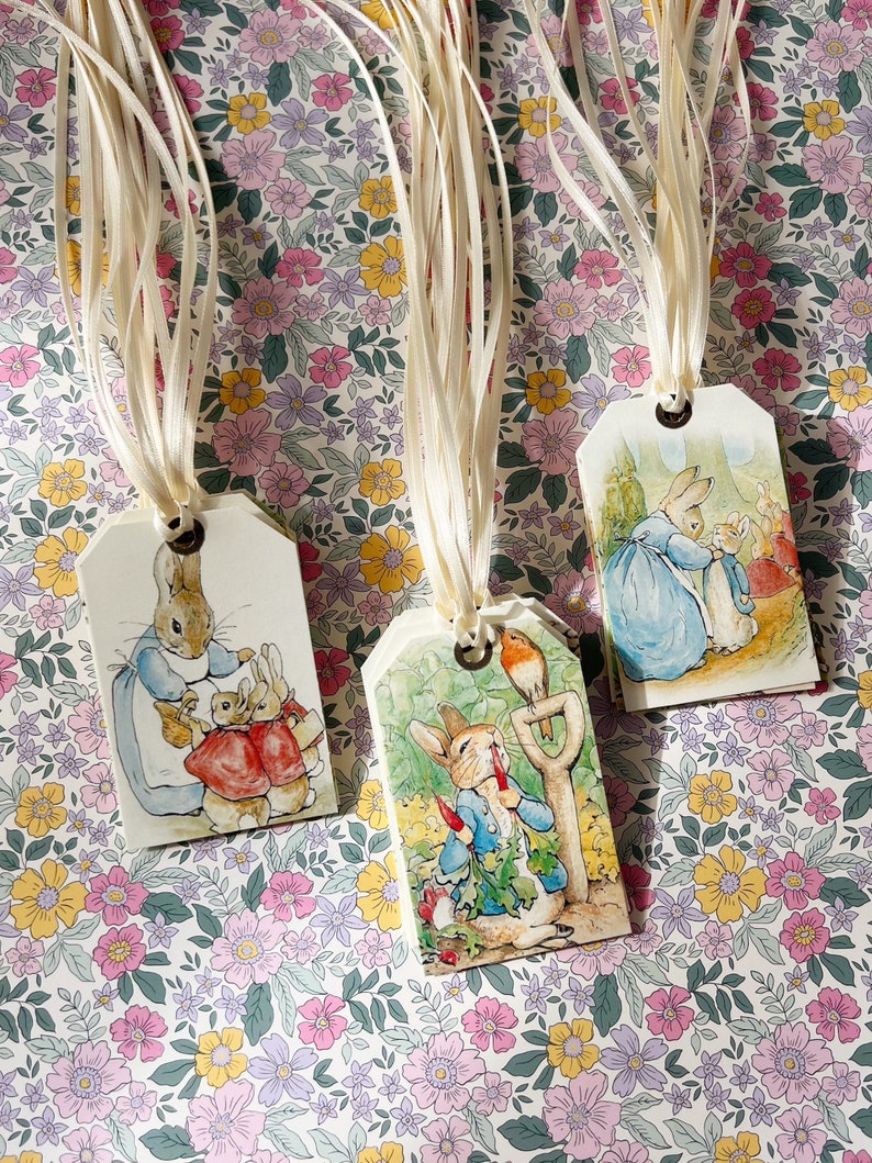 May include: Three Peter Rabbit gift tags with white satin ribbon. The tags feature illustrations of Peter Rabbit in different scenes. One tag shows Peter Rabbit with two other rabbits, another shows Peter Rabbit with a bird, and the last tag shows Peter Rabbit with a carrot.