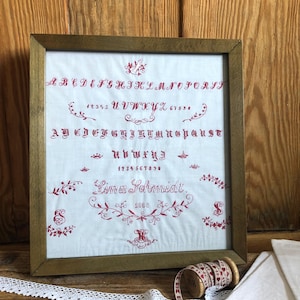 Puede incluir: Muestra de bordado enmarcada con un alfabeto rojo, números y elementos florales decorativos sobre tela blanca. El marco de madera rodea la muestra. Un carrete de cinta y encaje están en primer plano. Incluye el nombre "Lima Schmidt".