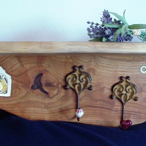 Könnte beinhalten: Ein Holzregal mit rustikalem, natürlichem Finish. Es verfügt über drei dekorative Haken mit verzierten Metalldesigns und eine kleine Keramikfliese mit einem Blumenmuster. Das Regal ist an einer weißen Wand montiert und hat einen Strauß Lavendelblüten obenauf.