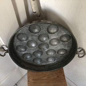 Op de afbeelding: Een vintage, ovale metalen pan met meerdere afgeronde inkepingen, waarschijnlijk om te koken. De pan heeft twee sierlijke handvatten en een doffe, zilvergrijze afwerking. De pan staat tegen een witte achtergrond en een houten oppervlak.