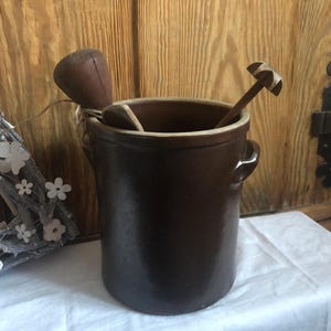 Pode incluir: Um pote de cerâmica marrom com uma abertura larga e uma alça de cada lado. O pote está cheio de utensílios de cozinha de madeira, incluindo uma colher de pau e um batedor de arame de madeira.