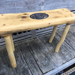 May include: A small wooden bench featuring a natural light-colored wood grain top with a live edge and a dark, oval-shaped metal inlay depicting a rose. The bench is supported by four cylindrical, light-colored wooden legs with a smooth finish.