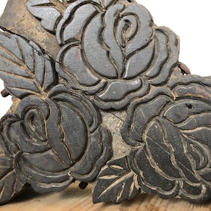 Könnte beinhalten: Nahaufnahme einer dunklen, verwitterten Holzschnitzerei mit drei detaillierten Rosenblüten und Blättern. Die Schnitzerei hat ein rustikales, antikes Aussehen mit sichtbarer Holzmaserung und Textur, was darauf hindeutet, dass es sich um ein altes Dekorationsobjekt handelt.
