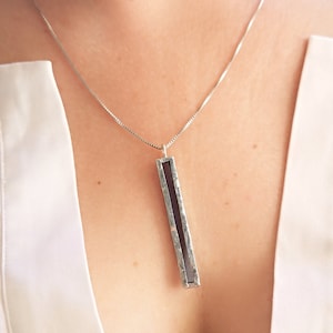 Peut inclure: Un collier en chaîne argent avec un pendentif rectangulaire long. Le pendentif est en argent et contient un morceau de verre rectangulaire gris foncé à l'intérieur.