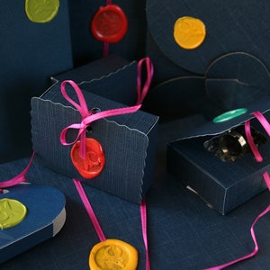Op de afbeelding: Marineblauwe geschenkdozen en enveloppen met kleurrijke zegellakken en roze linten. De dozen zijn in verschillende vormen en maten, sommige met geschulpte randen. De zegellakken zijn in tinten geel, rood en groen.
