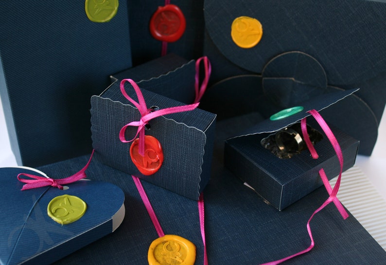 Op de afbeelding: Marineblauwe geschenkdozen en enveloppen met kleurrijke zegellakken en roze linten. De dozen zijn in verschillende vormen en maten, sommige met geschulpte randen. De zegellakken zijn in tinten geel, rood en groen.