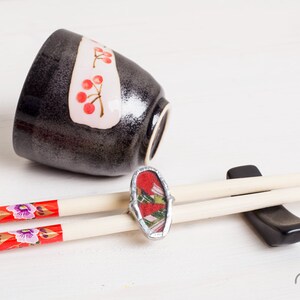 May include: A pair of chopsticks with red and purple floral designs on the ends, resting on a black chopstick rest. A ring with a red and green patterned design is on top of the chopsticks. A black bowl with a white band and red cherry designs is in the background.