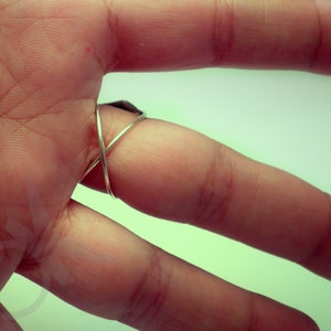 Op de afbeelding: Een zilverkleurige ring met een X-vormig ontwerp. De ring wordt om een vinger gedragen, waarbij de band over de bovenkant kruist. De ring lijkt gemaakt te zijn van een dunne metalen draad.