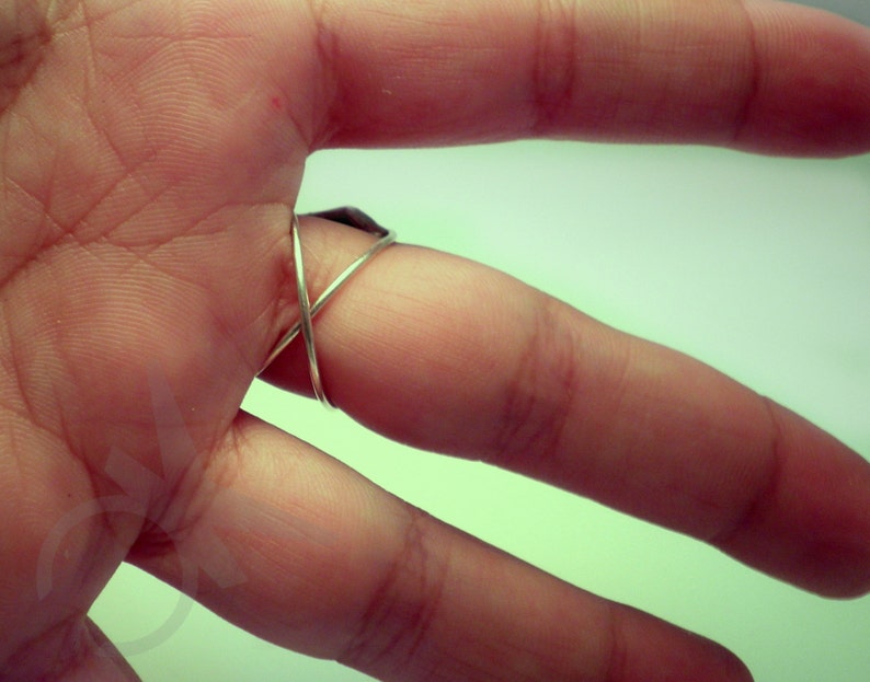 Op de afbeelding: Een zilverkleurige ring met een X-vormig ontwerp. De ring wordt om een vinger gedragen, waarbij de band over de bovenkant kruist. De ring lijkt gemaakt te zijn van een dunne metalen draad.