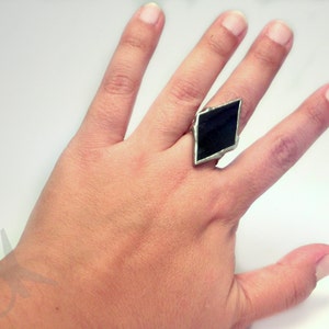 Op de afbeelding: Een zilverkleurige ring met een grote, donkere, ruitvormige steen. De zetting van de ring lijkt van metaal te zijn en de steen heeft een diepe, effen kleur. De ring wordt op een vinger gedragen, tegen een witte achtergrond.
