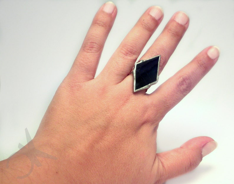 Op de afbeelding: Een zilverkleurige ring met een grote, donkere, ruitvormige steen. De zetting van de ring lijkt van metaal te zijn en de steen heeft een diepe, effen kleur. De ring wordt op een vinger gedragen, tegen een witte achtergrond.