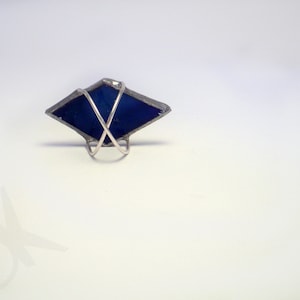 Op de afbeelding: Handgemaakte ring met een donkerblauw, ruitvormig glaselement. Het glas is ingekapseld in een zilverkleurig metalen frame en gemonteerd op een eenvoudige ringband. Het ontwerp is geometrisch en modern.