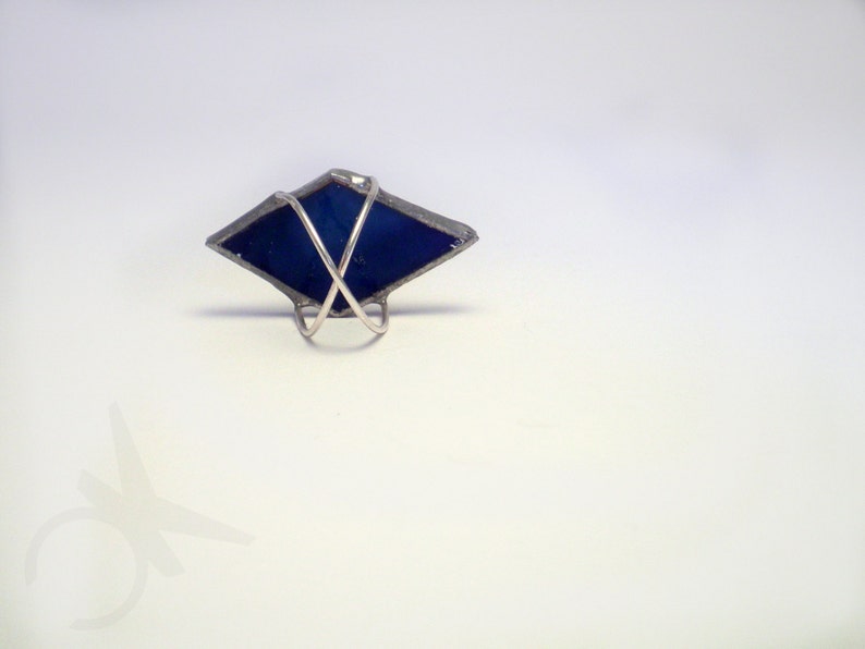 Op de afbeelding: Handgemaakte ring met een donkerblauw, ruitvormig glaselement. Het glas is ingekapseld in een zilverkleurig metalen frame en gemonteerd op een eenvoudige ringband. Het ontwerp is geometrisch en modern.