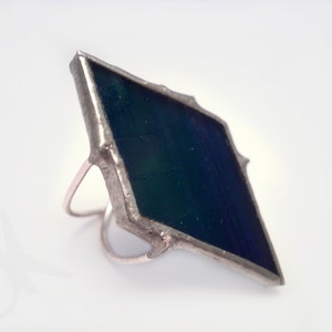Op de afbeelding: Een zilverkleurige ring met een groot, ruitvormig glas-in-lood element. Het glas heeft een donkerblauwe en groene kleurovergang. De ring heeft een dunne, eenvoudige band, die het gedurfde glasontwerp aanvult.