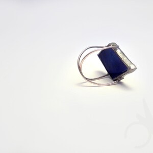 Op de afbeelding: Een zilverkleurige ring met een vierkante, diepblauwe edelsteen. De band van de ring heeft een dubbele lus, en de edelsteen is gezet in een zilveren metalen frame. De ring wordt gepresenteerd op een witte ondergrond.