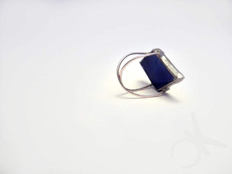 Op de afbeelding: Een zilverkleurige ring met een vierkante, diepblauwe edelsteen. De band van de ring heeft een dubbele lus, en de edelsteen is gezet in een zilveren metalen frame. De ring wordt gepresenteerd op een witte ondergrond.