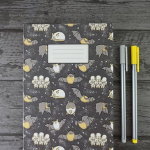 Op de afbeelding: Een notitieboek met een donkergrijze kaft met een patroon van cartoonvleermuizen in verschillende poses, met gele en witte accenten. Twee pennen, een grijze en een gele, liggen naast het notitieboek op een houten oppervlak.