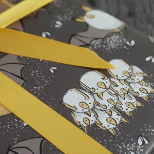 Op de afbeelding: Cadeau verpakt met grijs papier met een vleermuismotief en een felgeel lint. Het vleermuisontwerp heeft witte en gele accenten. Het lint is in een strik geknoopt.