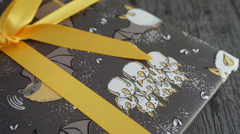 Op de afbeelding: Cadeau verpakt met grijs papier met een vleermuismotief en een felgeel lint. Het vleermuisontwerp heeft witte en gele accenten. Het lint is in een strik geknoopt.