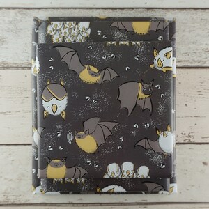 Op de afbeelding: Twee op elkaar gestapelde cadeauverpakkingen verpakt in donkergrijs papier met een herhaald patroon van cartoon vleermuizen in grijs en geel. De vleermuizen staan in verschillende poses, met kleine witte spikkels.