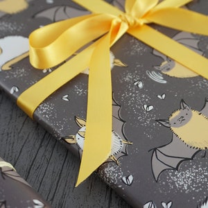 Op de afbeelding: Cadeau verpakt met grijs cadeaupapier met een patroon van gele en witte cartoonvleermuizen. Het cadeau is vastgebonden met een felgeel satijnen lint. Een rol bijpassend cadeaupapier staat op de voorgrond.