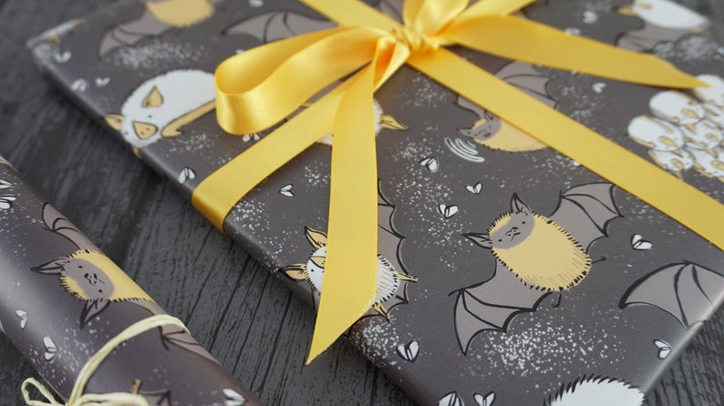Op de afbeelding: Cadeau verpakt met grijs cadeaupapier met een patroon van gele en witte cartoonvleermuizen. Het cadeau is vastgebonden met een felgeel satijnen lint. Een rol bijpassend cadeaupapier staat op de voorgrond.
