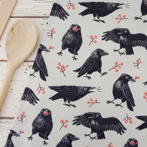 May include: A light grey kitchen towel featuring a repeating pattern of black crows in various poses, some with wings spread or beaks open, interspersed with small clusters of orange-red berries. A wooden spoon is positioned along the left edge.