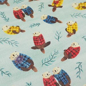 May include: Watercolor illustration featuring otters wearing plaid shirts in red, blue, and yellow. The otters are scattered across a light blue background with green leafy sprigs. The design has a whimsical, playful feel.