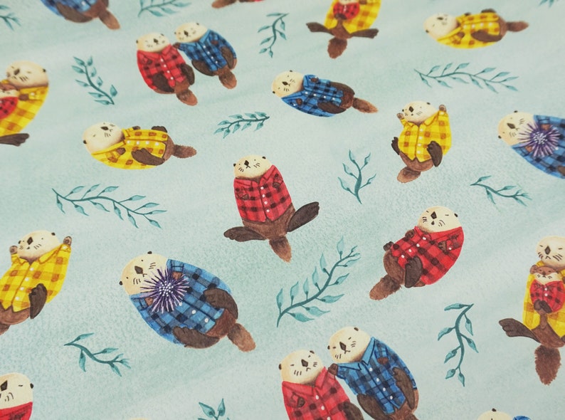 May include: Watercolor illustration featuring otters wearing plaid shirts in red, blue, and yellow. The otters are scattered across a light blue background with green leafy sprigs. The design has a whimsical, playful feel.