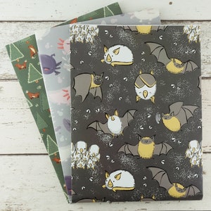 Op de afbeelding: Drie rechthoekige notitieboekjes met ge&iuml;llustreerde covers. De bovenste cover heeft een donkergrijze achtergrond met witte en gele cartoonvleermuizen. De middelste cover toont lichtblauwe axolotls. De onderste cover is groen met kleine oranje vossen en bomen.