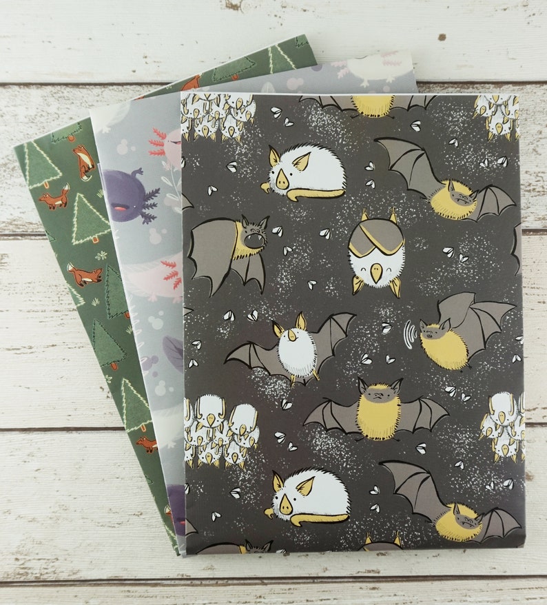Op de afbeelding: Drie rechthoekige notitieboekjes met ge&iuml;llustreerde covers. De bovenste cover heeft een donkergrijze achtergrond met witte en gele cartoonvleermuizen. De middelste cover toont lichtblauwe axolotls. De onderste cover is groen met kleine oranje vossen en bomen.