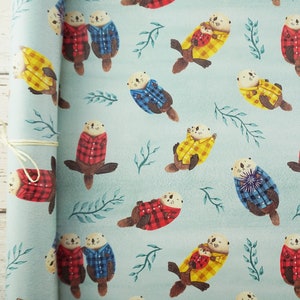 May include: A roll of light blue wrapping paper featuring a repeating pattern of otters wearing plaid shirts in red, blue, and yellow. The otters are interspersed with green leaf sprigs. The paper is tied with a natural twine.