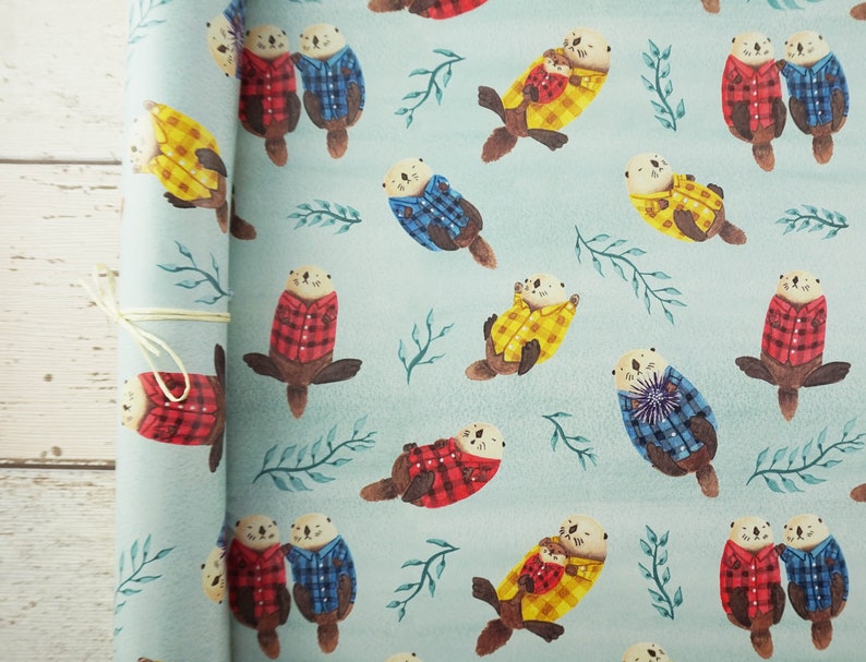 May include: A roll of light blue wrapping paper featuring a repeating pattern of otters wearing plaid shirts in red, blue, and yellow. The otters are interspersed with green leaf sprigs. The paper is tied with a natural twine.