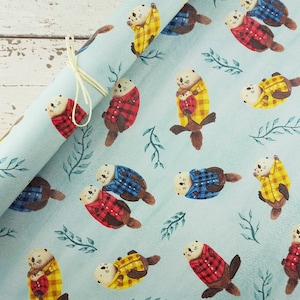 May include: Light blue wrapping paper featuring a repeating pattern of watercolor otters wearing plaid shirts in red, yellow, and blue. The otters are interspersed with green leafy sprigs. A cream-colored ribbon is tied around the rolled paper.