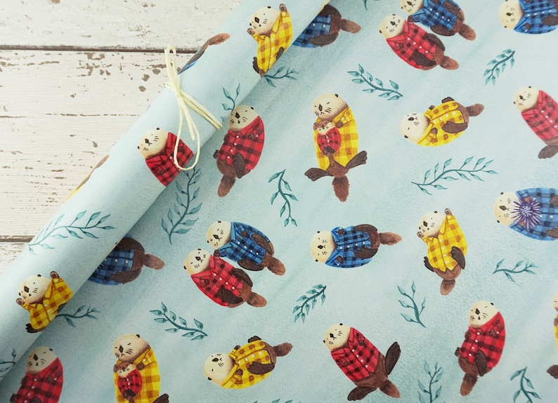 May include: Light blue wrapping paper featuring a repeating pattern of watercolor otters wearing plaid shirts in red, yellow, and blue. The otters are interspersed with green leafy sprigs. A cream-colored ribbon is tied around the rolled paper.