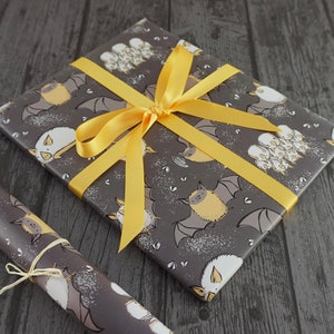 Op de afbeelding: Een ingepakt cadeau met grijs papier met vleermuismotief en een felgeel lint. Het papier bevat illustraties van vleermuizen in verschillende poses, met gele en witte accenten. Een bijpassende rol papier is vastgebonden met touw.