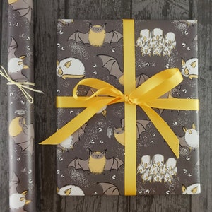 Op de afbeelding: Een ingepakt cadeau en een rol cadeaupapier, beide versierd met een herhaald patroon van cartoonvleermuizen in grijstinten, beige en wit. Het cadeau is vastgebonden met een felgeel lint. Het cadeaupapier heeft een bijpassend vleermuisontwerp.