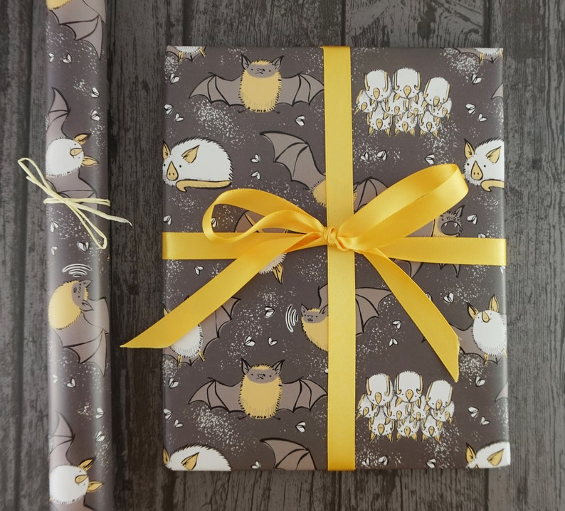 Op de afbeelding: Een ingepakt cadeau en een rol cadeaupapier, beide versierd met een herhaald patroon van cartoonvleermuizen in grijstinten, beige en wit. Het cadeau is vastgebonden met een felgeel lint. Het cadeaupapier heeft een bijpassend vleermuisontwerp.