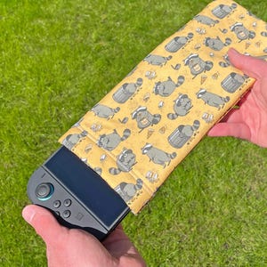 May include: A yellow fabric case featuring a repeating pattern of cartoon raccoons, bins, and pizza slices. The case holds a black handheld games console. The case is held by a person.