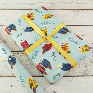 May include: Gift wrap featuring a repeating pattern of otters wearing plaid jackets in red, yellow, and blue on a light blue background. The wrapped gift is tied with a yellow ribbon. A roll of matching paper is next to the gift.