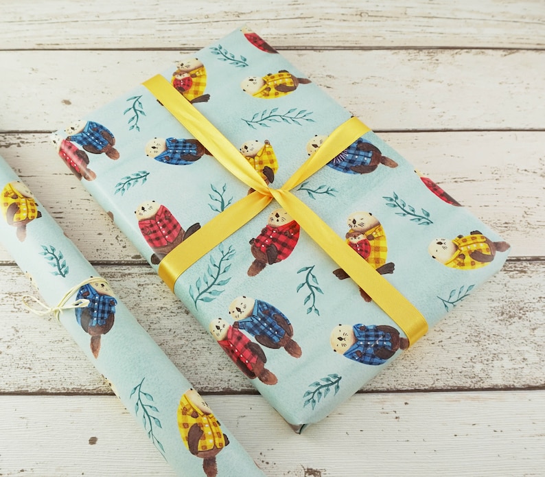 May include: Gift wrap featuring a repeating pattern of otters wearing plaid jackets in red, yellow, and blue on a light blue background. The wrapped gift is tied with a yellow ribbon. A roll of matching paper is next to the gift.