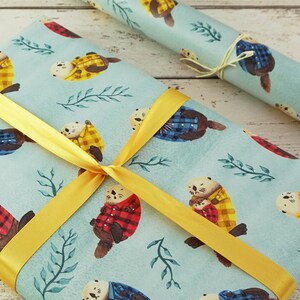 May include: Gift wrap featuring a light blue background with watercolor otters wearing plaid jackets in red, yellow, and blue. The wrapped gift is tied with a yellow ribbon. A roll of matching wrapping paper is also visible.