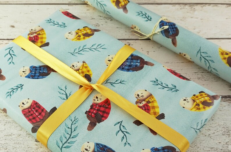 May include: Gift wrap featuring a light blue background with watercolor otters wearing plaid jackets in red, yellow, and blue. The wrapped gift is tied with a yellow ribbon. A roll of matching wrapping paper is also visible.