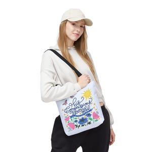 May include: A white tote bag with a black shoulder strap featuring a colourful floral design, a yellow sun, a butterfly, and the text His compassions never fail they are new every morning in blue and black script lettering on the front.