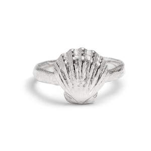 May include: A silver ring featuring a central scallop shell motif with a textured, organic band. The metal has a polished finish that highlights the vertical ridges and fan-like shape of the shell, creating a detailed and reflective surface.