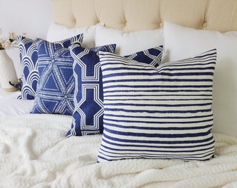 May include: A collection of four square throw pillows featuring various navy blue and white patterns, including horizontal stripes, geometric shapes, and arched lines, arranged in a row against a neutral-colored tufted headboard.